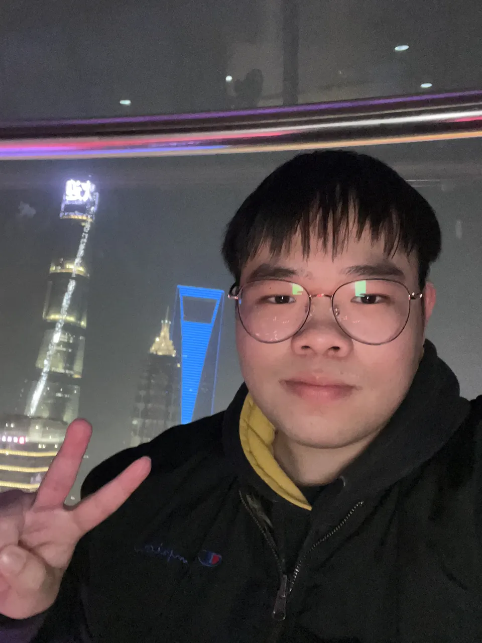 Jinhao making a peace sign at night, the lit Lujiazui towers across the river