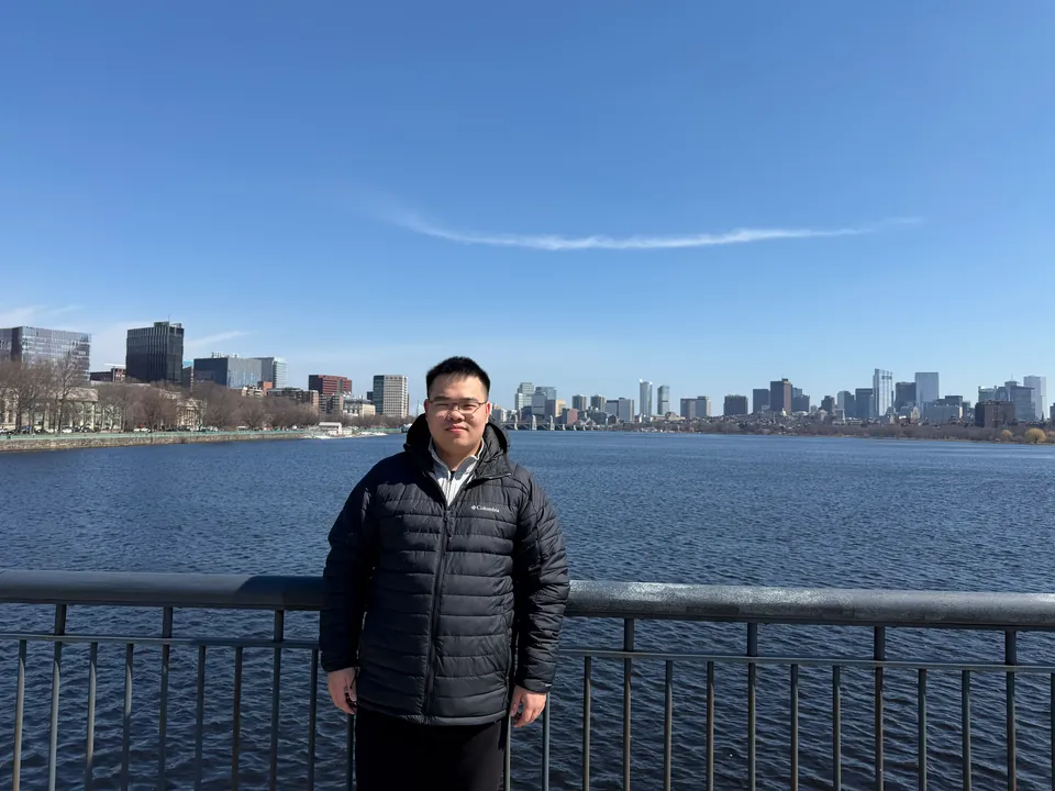 Jinhao by a railing on the Charles River with the Boston skyline behind