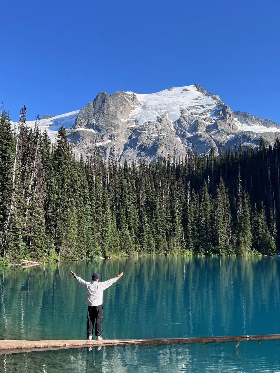 Jinhao with arms open on a log in a turquoise lake below a glacier