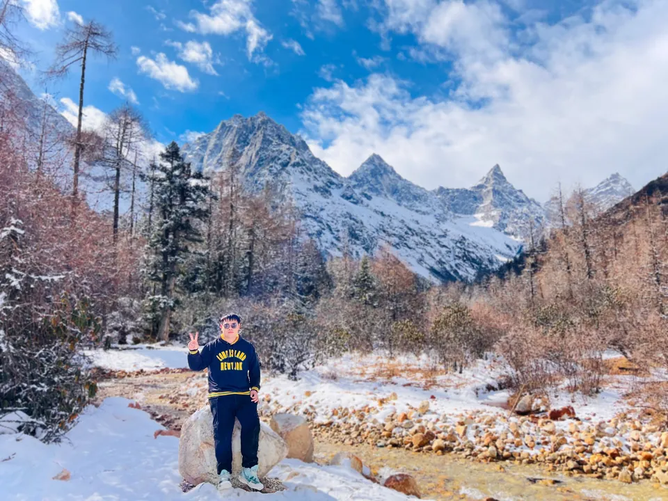 Jinhao making a peace sign on a rock by a frozen stream, snowy peaks behind