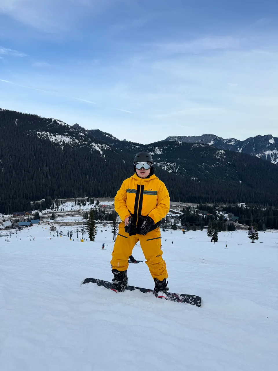 Jinhao in a yellow snow suit standing on a snowboard, forested hills behind