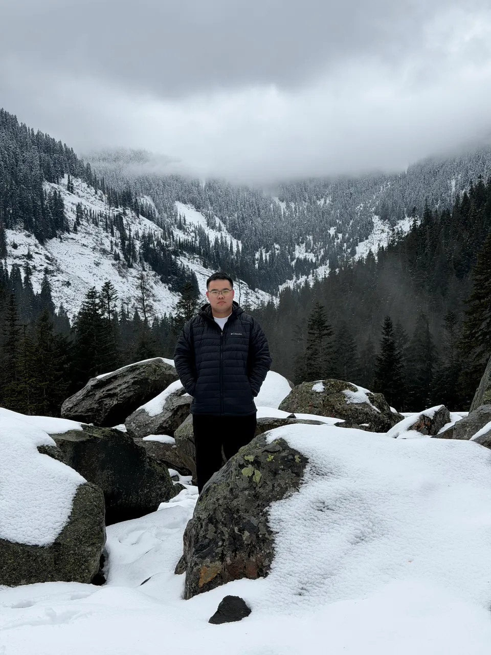 Jinhao among snowy boulders, a forested valley in low cloud behind